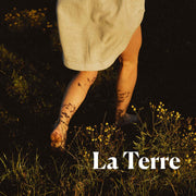 Photo de couverture pour le parfum naturel La Terre de Floratropia. Femme qui marche dans un champ, vue de dos, centré sur ses jambes.
